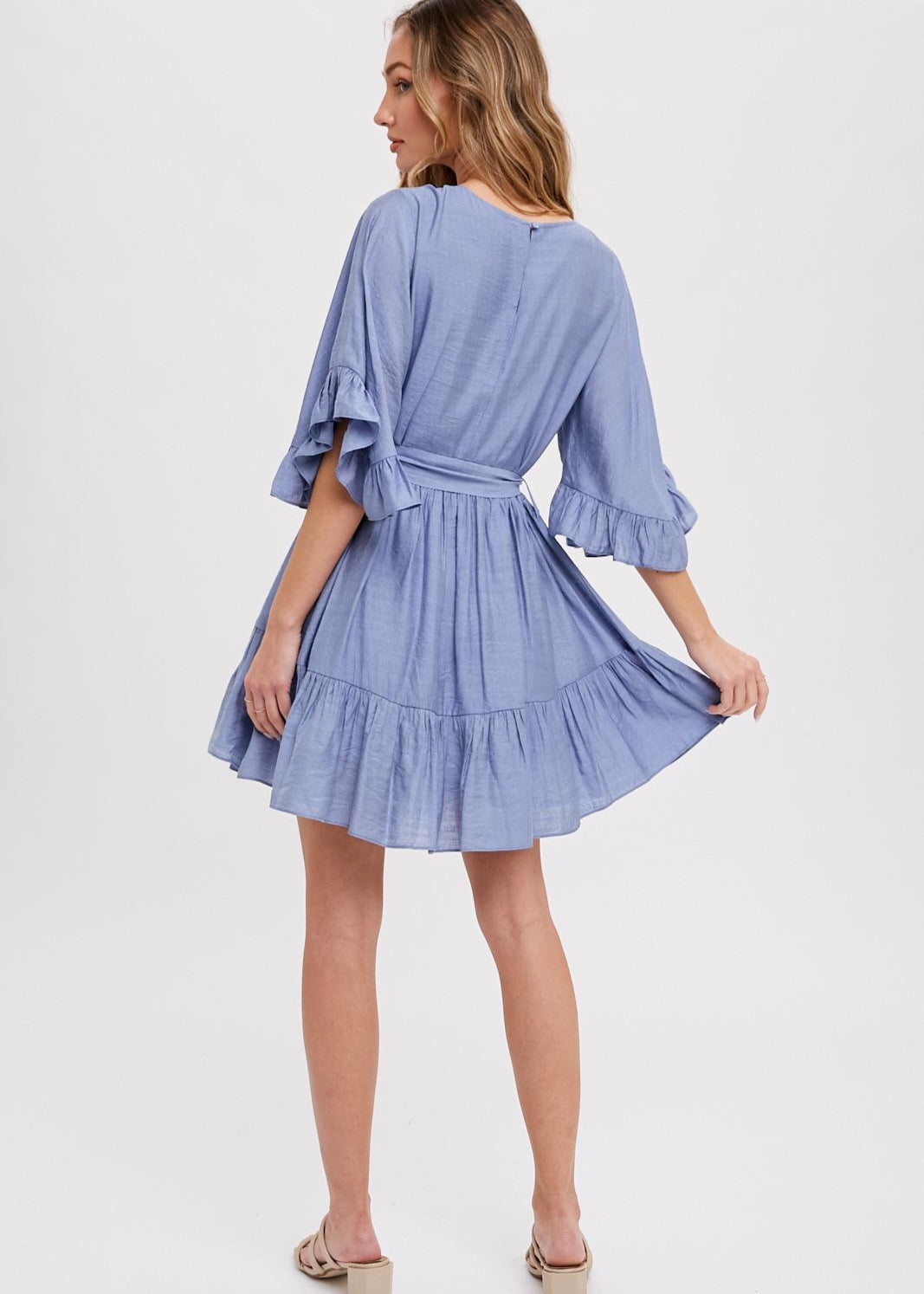 Chambray mini dress features a round neck, along with flounce sleeves that have a loose and flowy appearance. It also has a self-tie belt at the waist, which allows for a customizable and fitted look. The bottom of the garment is finished with ruffles and frill detailing, adding a decorative touch and creating a playful and feminine style.