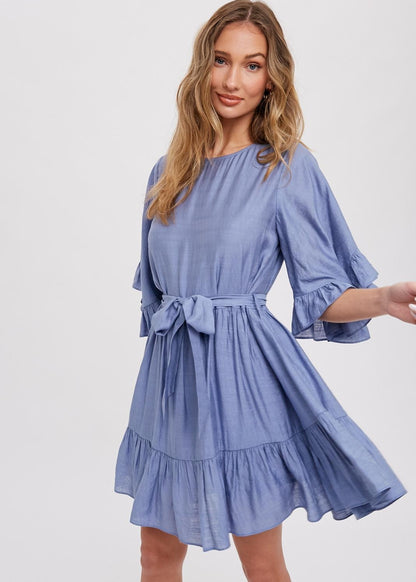 Chambray mini dress features a round neck, along with flounce sleeves that have a loose and flowy appearance. It also has a self-tie belt at the waist, which allows for a customizable and fitted look. The bottom of the garment is finished with ruffles and frill detailing, adding a decorative touch and creating a playful and feminine style.