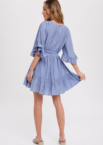 Chambray mini dress features a round neck, along with flounce sleeves that have a loose and flowy appearance. It also has a self-tie belt at the waist, which allows for a customizable and fitted look. The bottom of the garment is finished with ruffles and frill detailing, adding a decorative touch and creating a playful and feminine style.