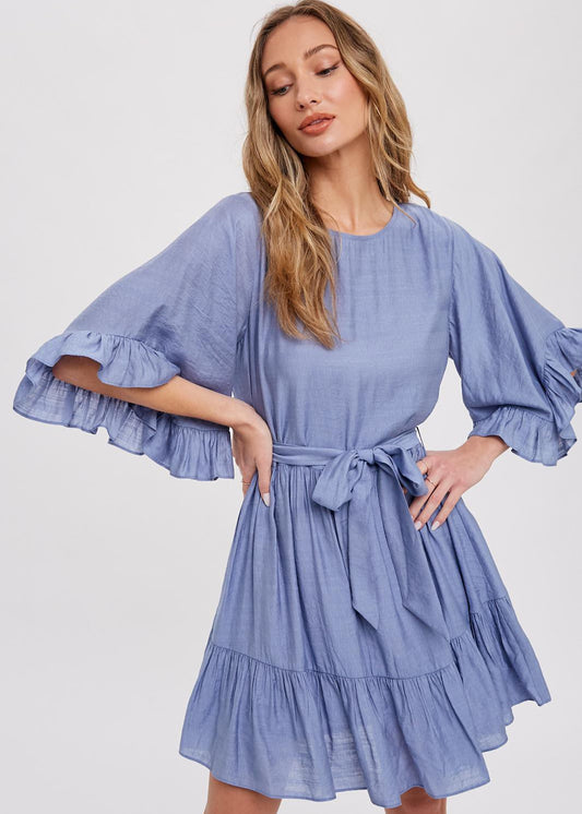 Chambray mini dress features a round neck, along with flounce sleeves that have a loose and flowy appearance. It also has a self-tie belt at the waist, which allows for a customizable and fitted look. The bottom of the garment is finished with ruffles and frill detailing, adding a decorative touch and creating a playful and feminine style.