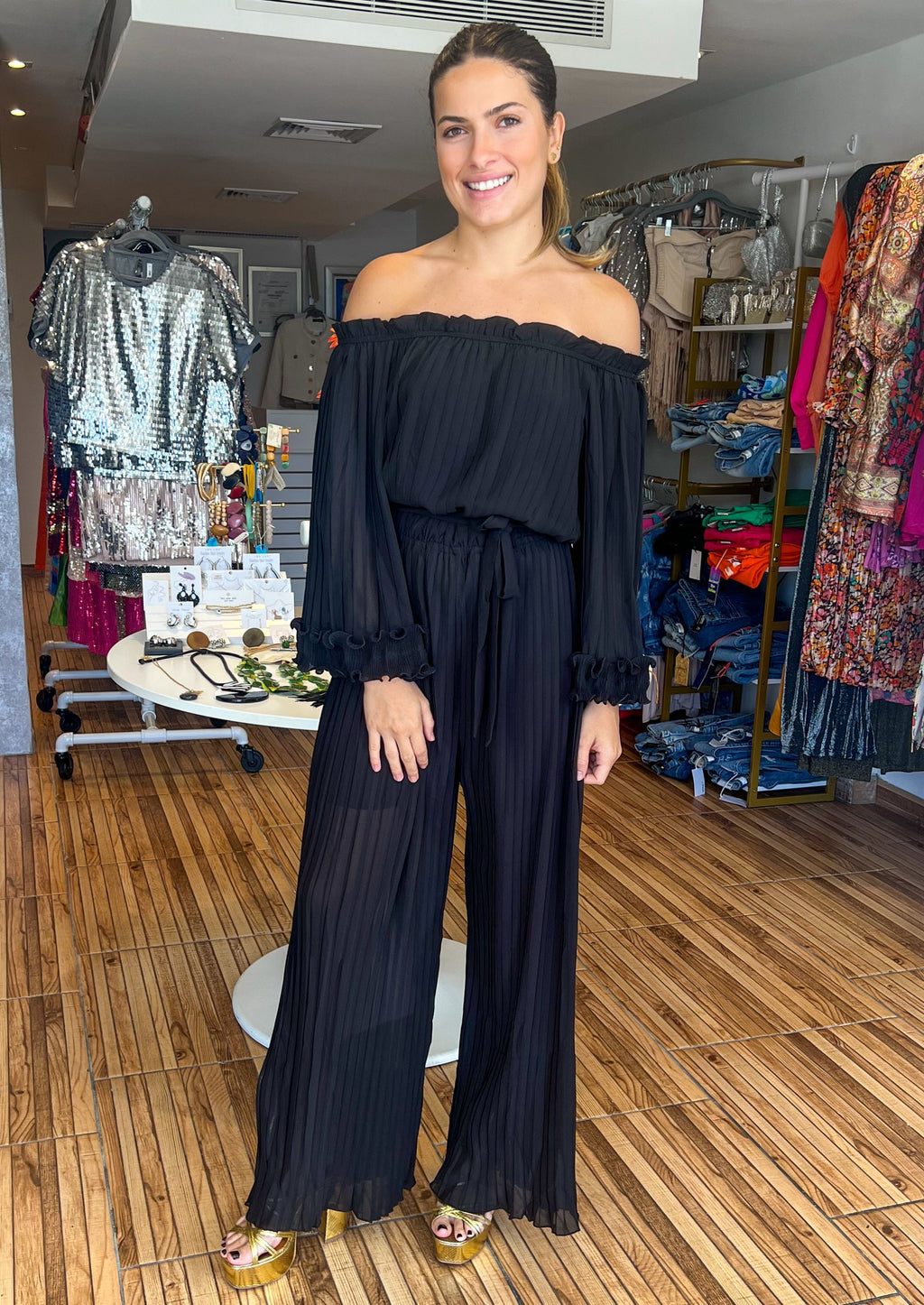 Black off shoulder pleated jumpsuit in chiffon fabric. Featuring elasticated off shoulder neck line, wide leg pants, lining, elastic at waist, long sleeves with ruffle lettuce edge detailing, and removable cinching waist tie, this jumpsuit is completely pleated and has a relaxed fit.